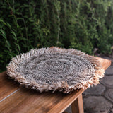 Round woven placemat on a wooden stool with a blurred green outdoor background