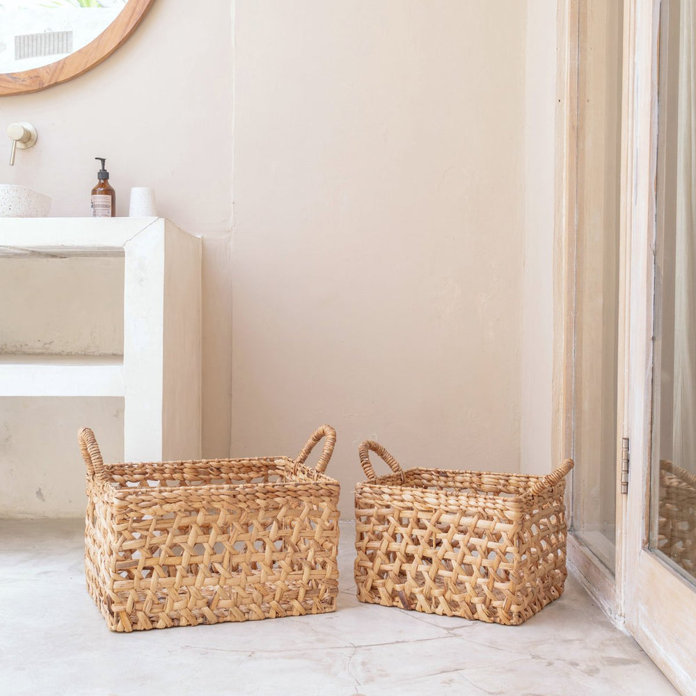 Two woven baskets on a light wooden floor with a neutral wall and a shelf in the background.
