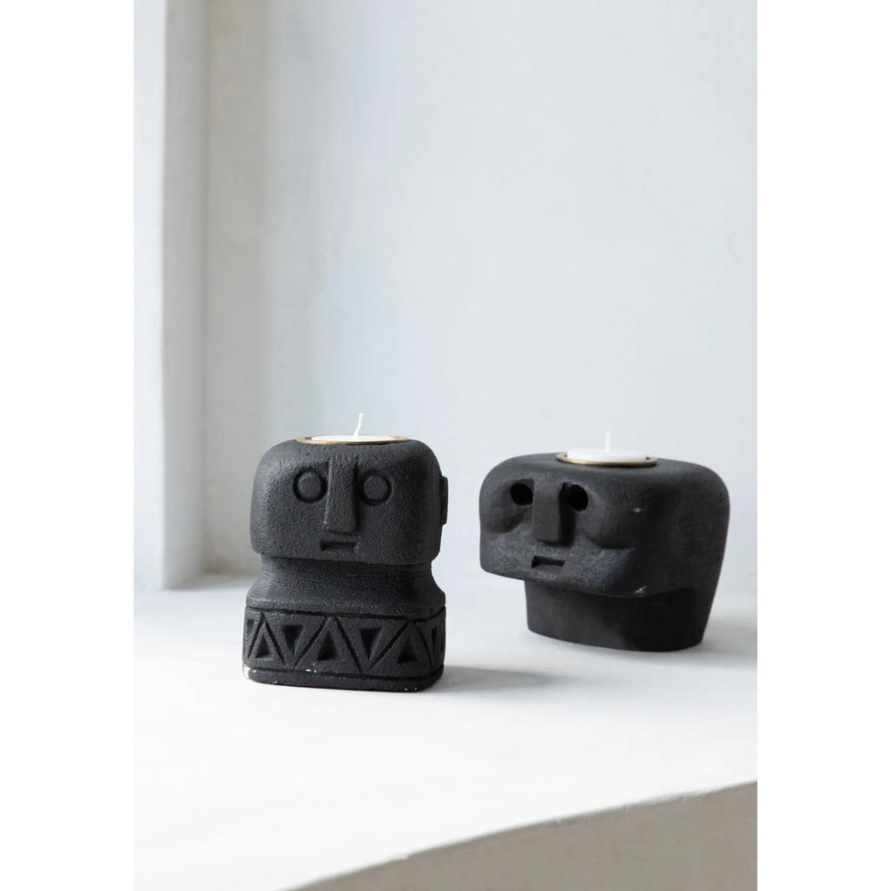 Two black ceramic face-shaped candle holders on a white surface with a light gray curtain background.