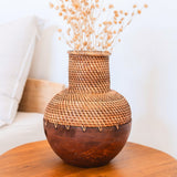Woven rattan and wooden vase with dried flowers on a wooden table.