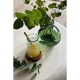 Glass jar with a straw and lemon slice on a wooden coaster, next to a green vase with eucalyptus leaves on a light surface.