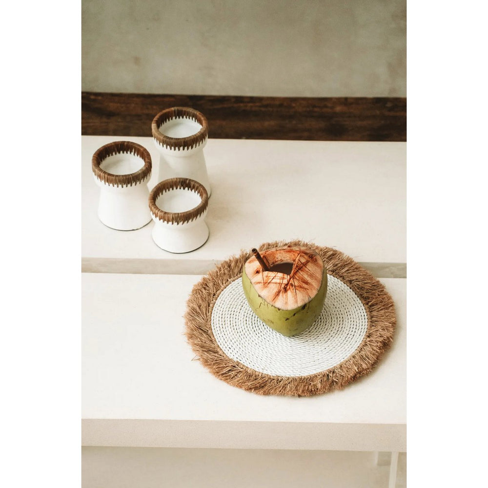 Coconut on a woven mat with three ceramic cups on a shelf in the background.