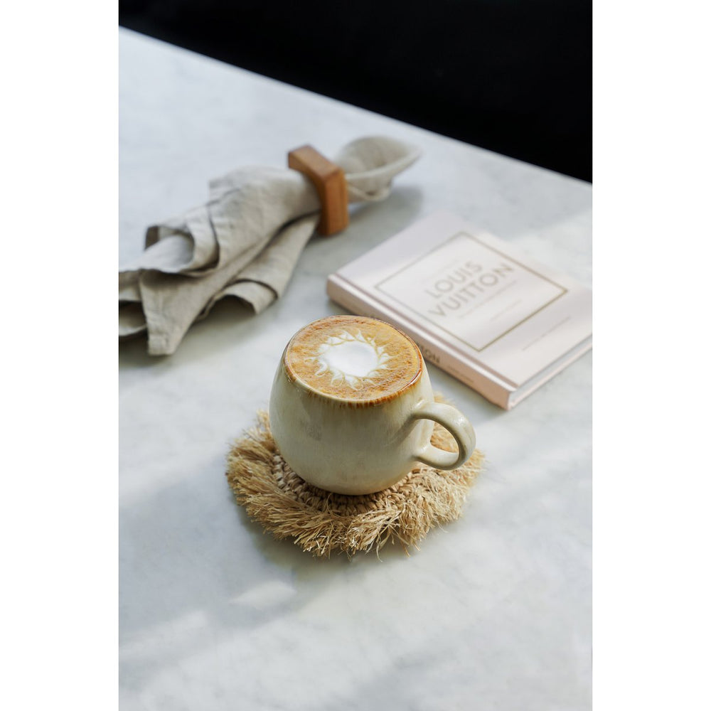 Cappuccino in a white cup on a straw coaster with a book and towel in the background.