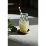 Glass jar with a straw on a wooden coaster, filled with a yellowish liquid, on a light surface.