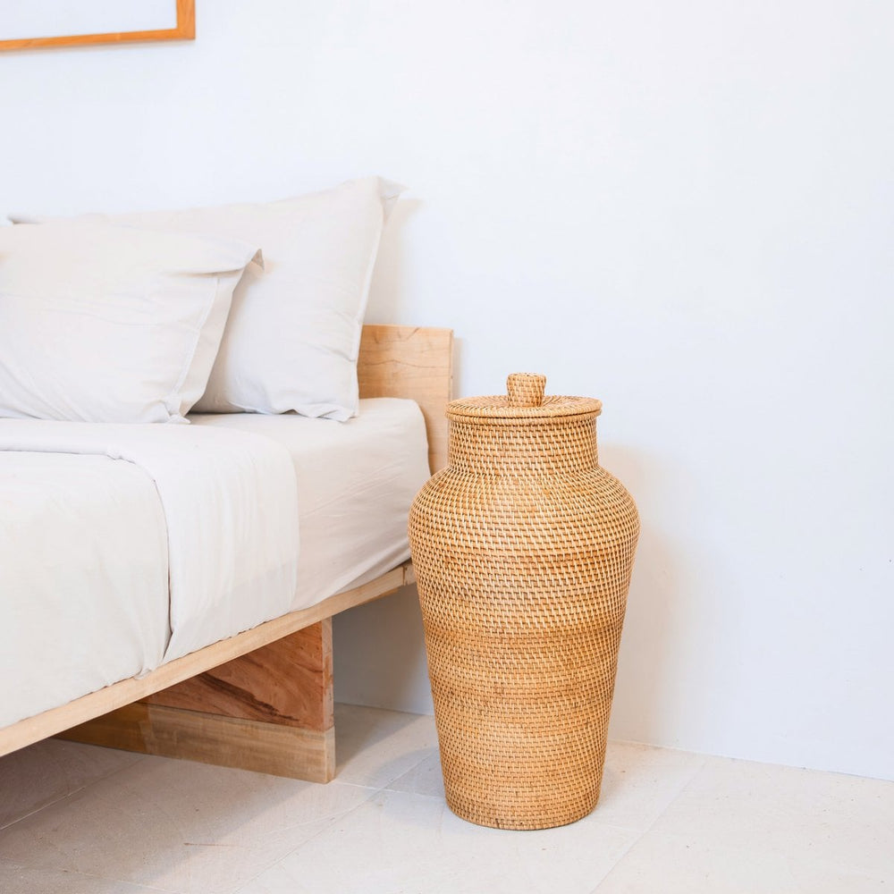 Woven rattan vase on a white surface next to a bed with white bedding.