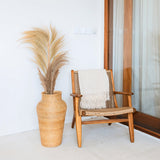 Wooden armchair with woven details next to a tall wicker vase with dried plants on a white floor.