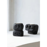 Two black ceramic face-shaped candle holders on a white surface with a light gray curtain background.