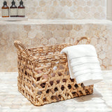 Woven basket with a white towel on a bathroom floor with tiled walls.
