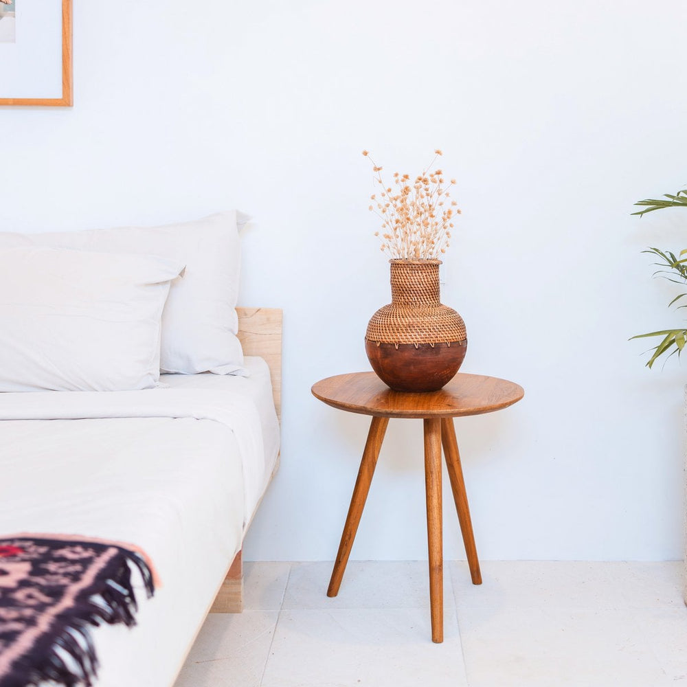 Wooden side table with a decorative vase next to a bed in a bedroom.