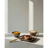Café setting with a cup of coffee, plate of cake, and bowl of fruit on a light surface.