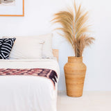 Wicker vase with dried pampas grass on a white surface next to a bed with patterned bedding.