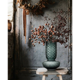 Decorative vase with dried flowers on a rustic wooden stool against a textured wall.