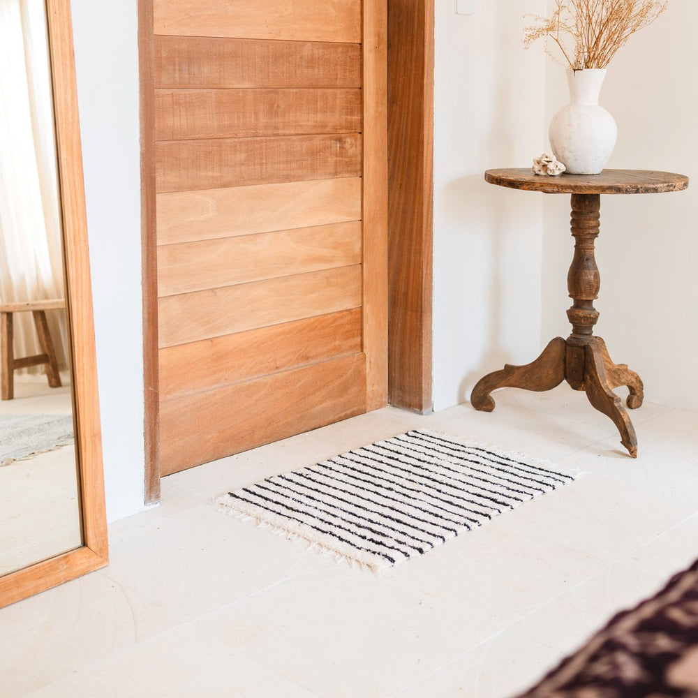 White rug with black stripes in front of a wooden door, with a wooden side table and vase in the background.