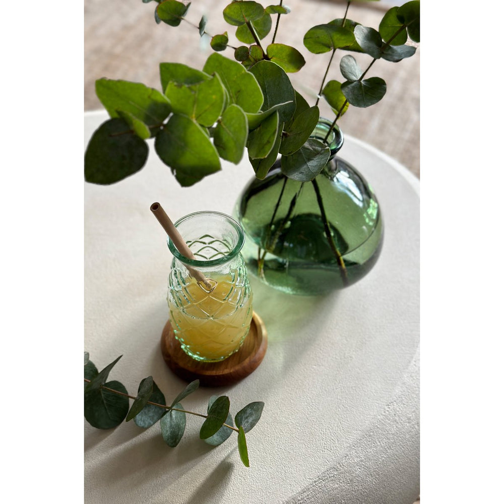 Glass jar with a straw and lemon slice on a wooden coaster, next to a green vase with eucalyptus leaves on a light surface.