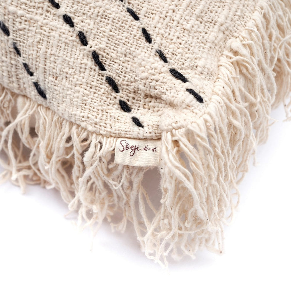 Beige woven blanket with black embroidery and fringes, featuring a brand label on a white background.