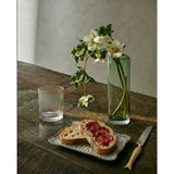 Bread with jam on a plate, knife, glass, and vase with flowers on a wooden table.