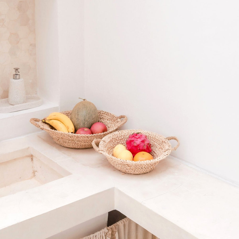 Two woven fruit bowls on a kitchen counter with fruits inside.