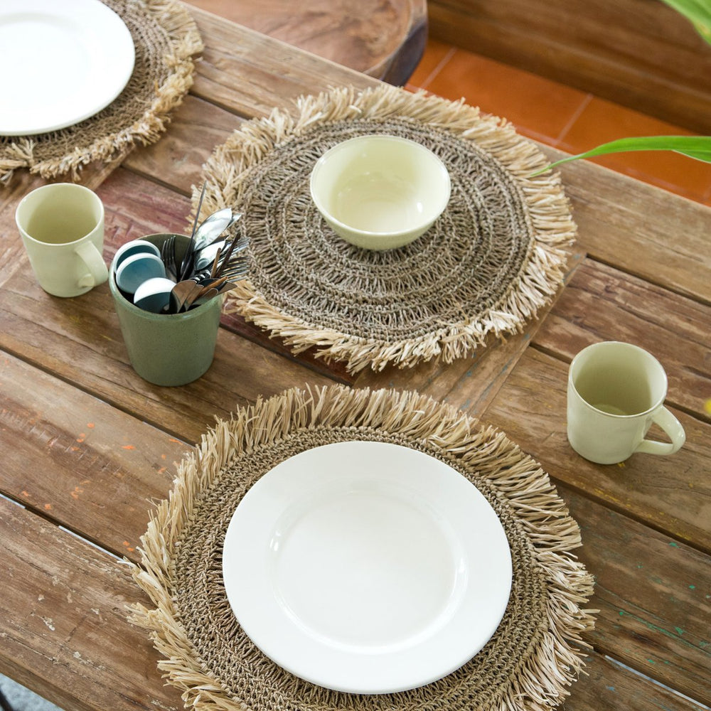 Table setting with woven placemats, white plates, and green mugs on a wooden table.